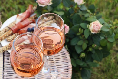Photo of Flowers near glasses of delicious rose wine and food on picnic basket outdoors, closeup