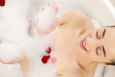 Photo of Happy woman taking bath in tub with foam and rose petals, top view
