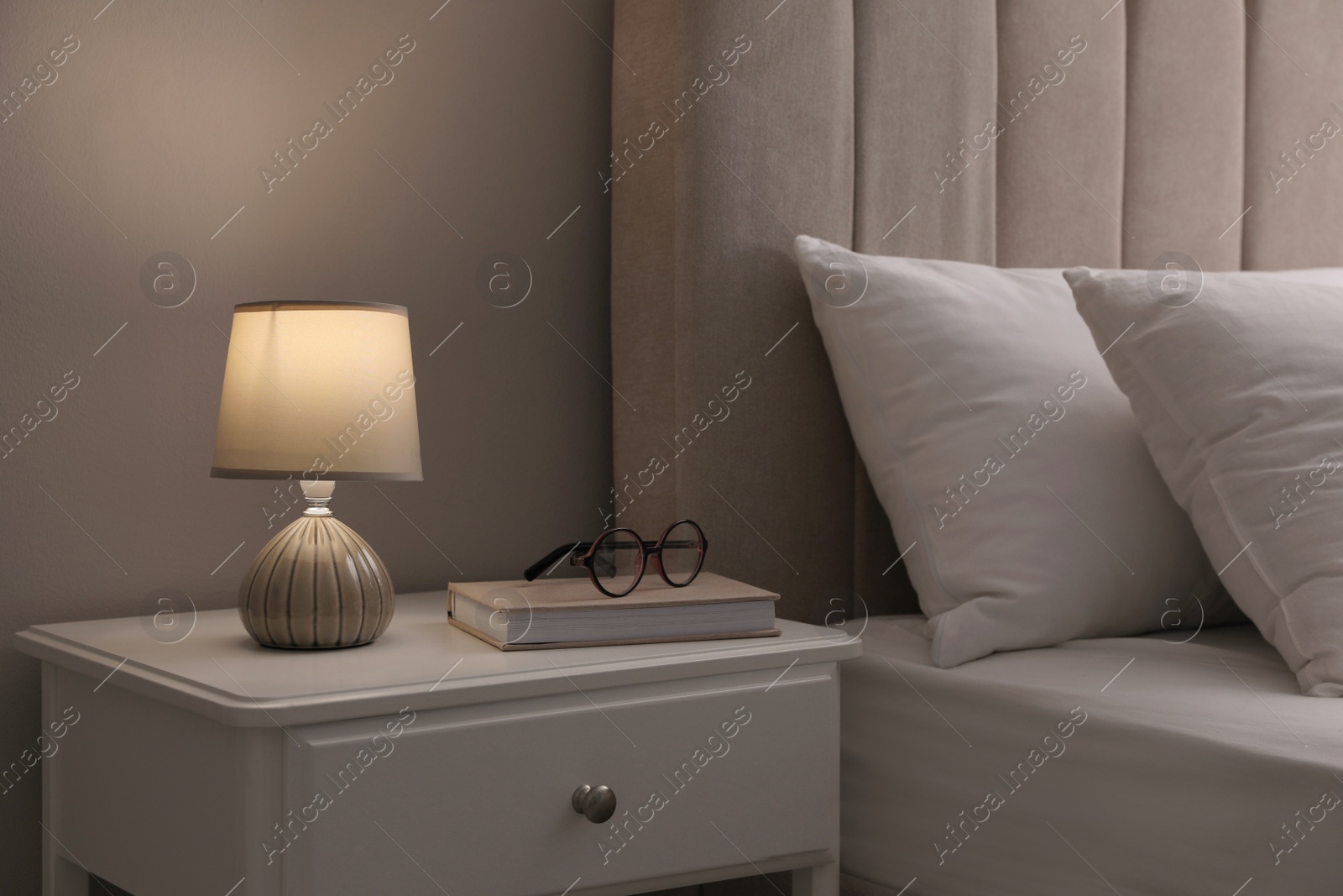 Photo of Stylish lamp, book and glasses on bedside table indoors. Bedroom interior elements