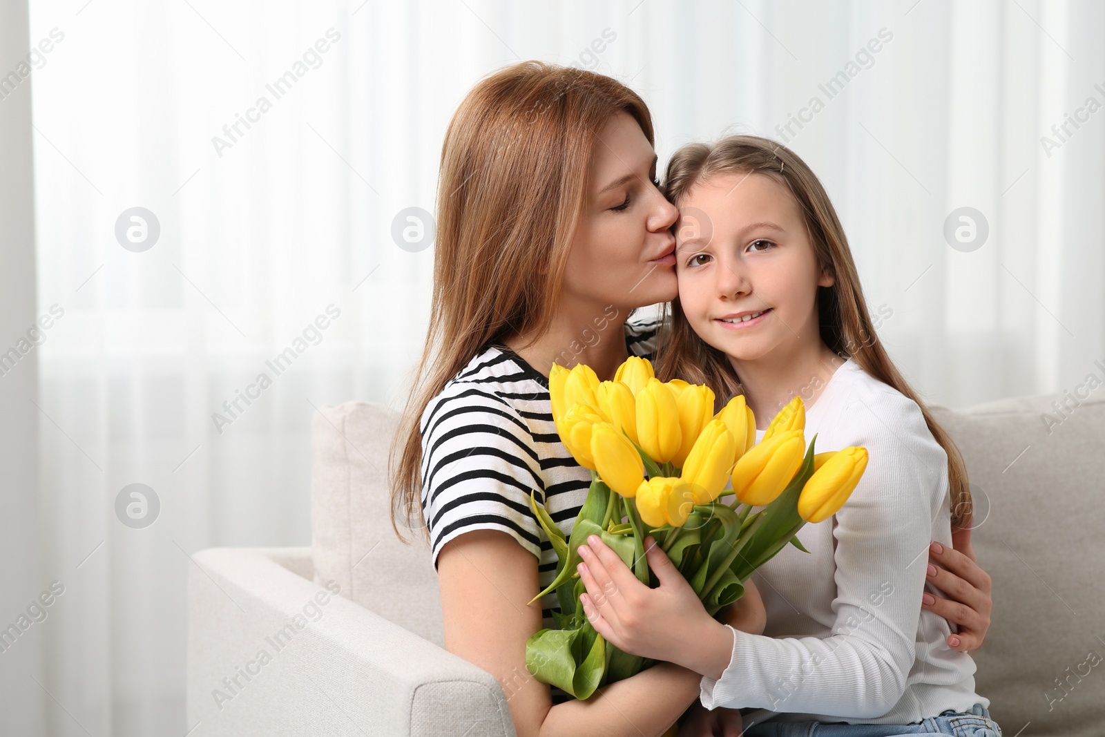 Photo of Mother and her cute daughter with bouquet of yellow tulips on sofa at home. Space for text