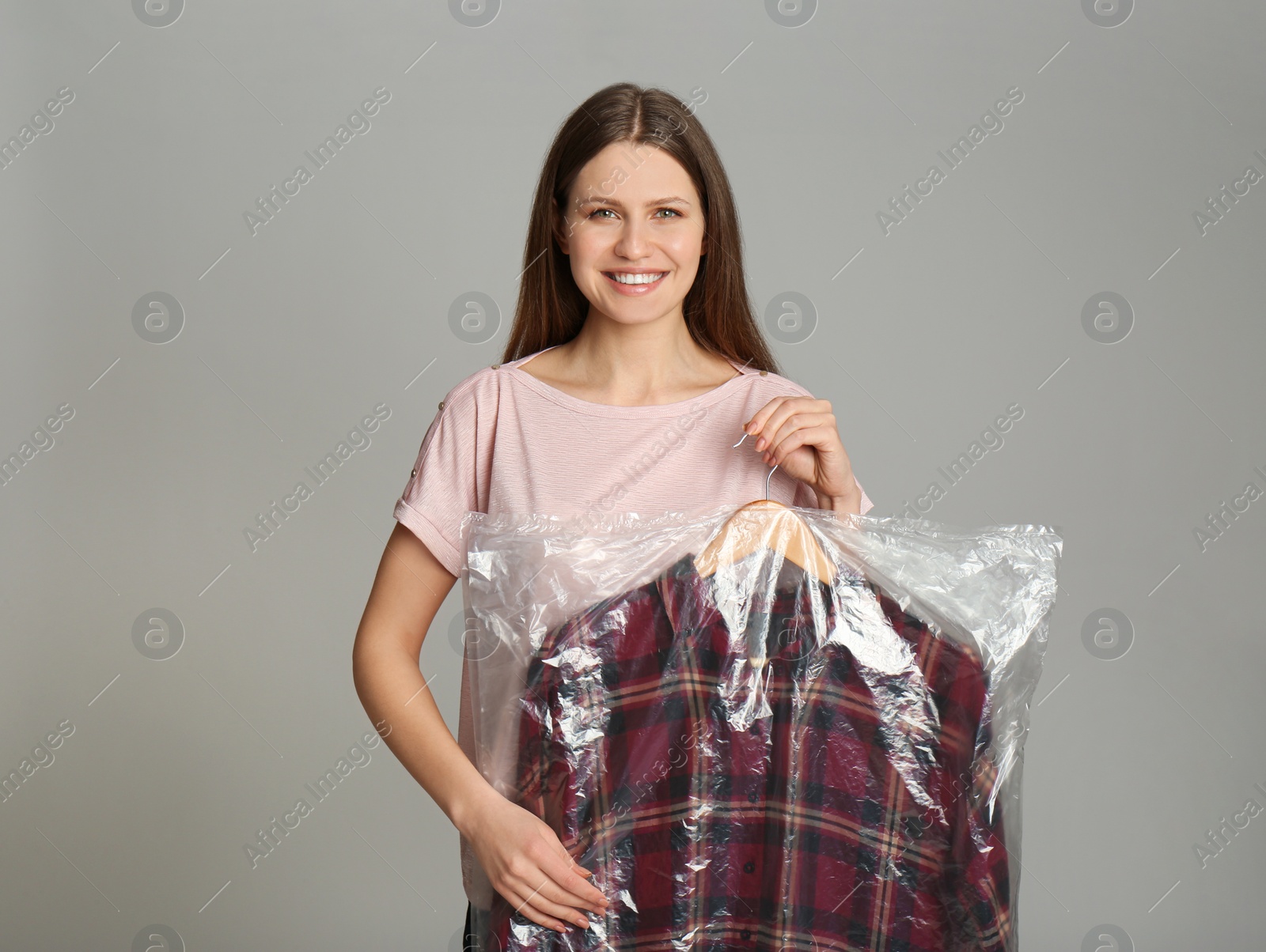 Photo of Young woman holding hanger with shirt in plastic bag on light grey background. Dry-cleaning service