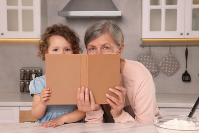 Photo of Cute little girl and her granny with recipe book in kitchen