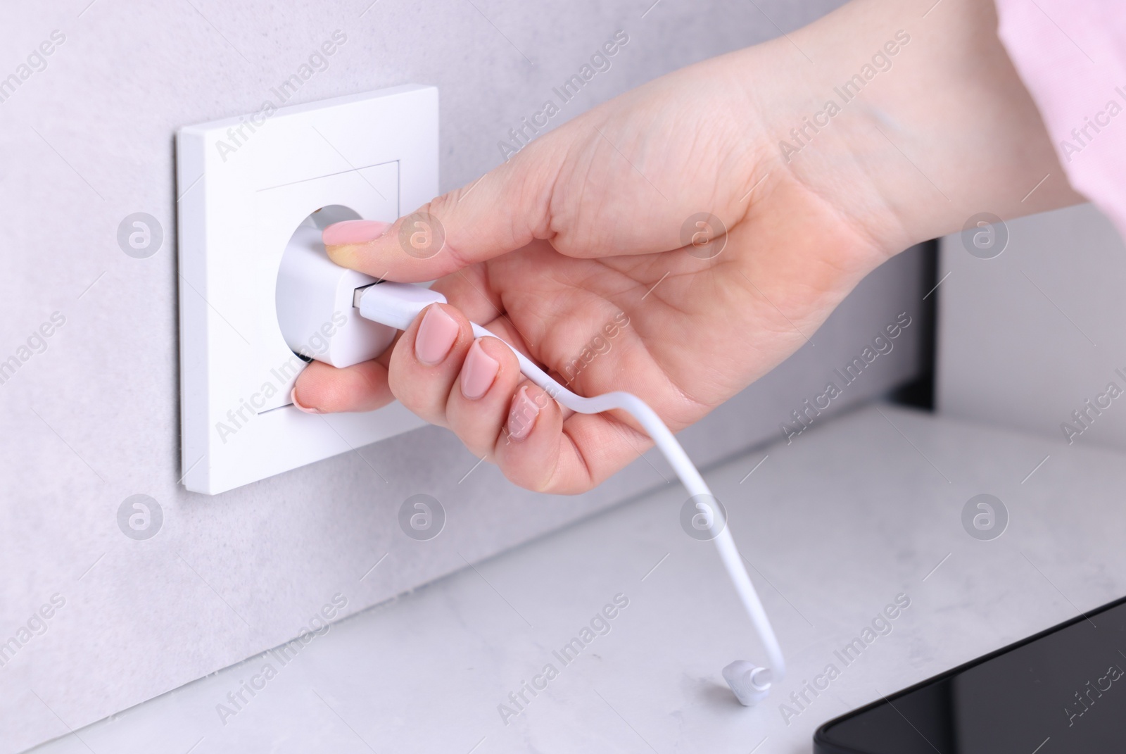 Photo of Woman plugging smartphone into power socket at white table indoors, closeup