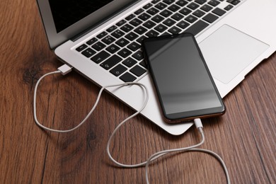 Photo of Smartphone connected with charge cable to laptop on wooden table. Space for text
