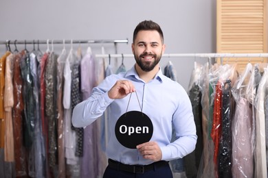 Photo of Dry-cleaning service. Happy worker holding Open sign near racks with clothes indoors