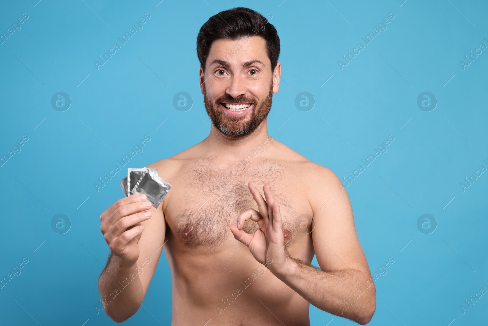 Photo of Naked man with condoms showing ok gesture on light blue background. Safe sex