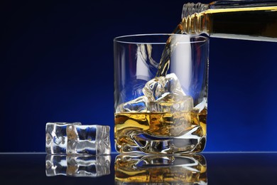 Photo of Pouring whiskey into glass with ice cubes at table against dark blue background