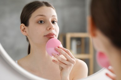 Photo of Washing face. Young woman with cleansing brush near mirror in bathroom