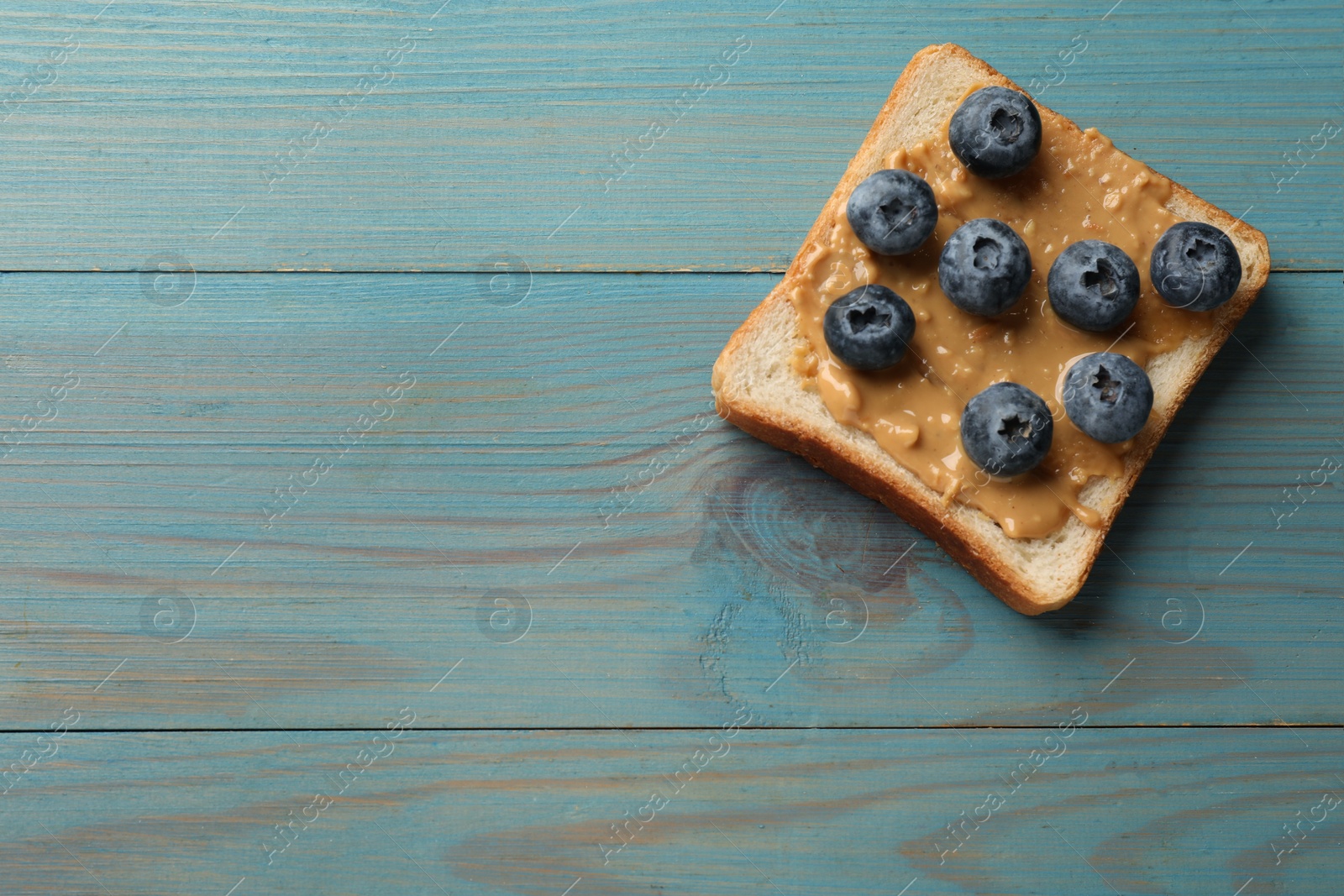 Photo of Delicious toast with peanut butter and blueberries on light blue wooden table, top view. Space for text