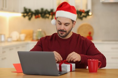 Photo of Celebrating Christmas online with exchanged by mail presents. Surprised man in Santa hat opening gift box during video call on laptop at home