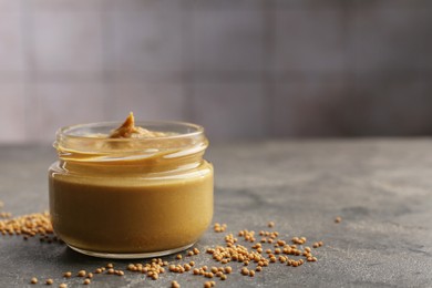 Photo of Tasty mustard sauce in jar and dry seeds on grey textured table, closeup. Space for text
