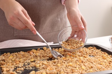 Photo of Woman putting granola from baking tray into glass bowl at table, closeup