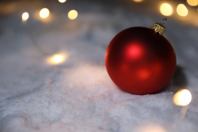 Photo of Christmas ball and fairy lights on white snow outdoors. Space for text