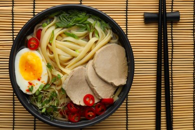 Photo of Delicious ramen with meat in bowl and chopsticks on bamboo mat, top view. Noodle soup