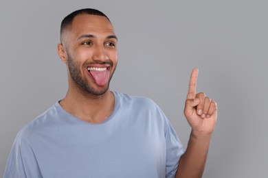Photo of Happy young man showing his tongue and pointing at something on light grey background