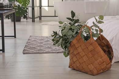 Photo of Stylish wicker basket with fresh eucalyptus branches and flowers on floor in bedroom