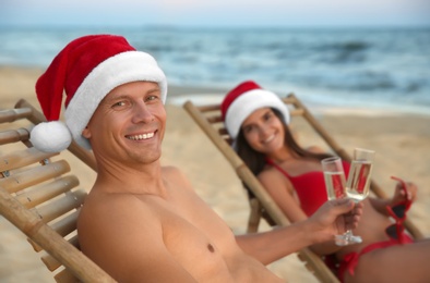 Photo of Happy couple wearing Santa hats and drinking champagne together on beach. Christmas vacation