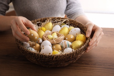Photo of Woman with basket full of dyed Easter eggs at wooden table, closeup. Easter celebration