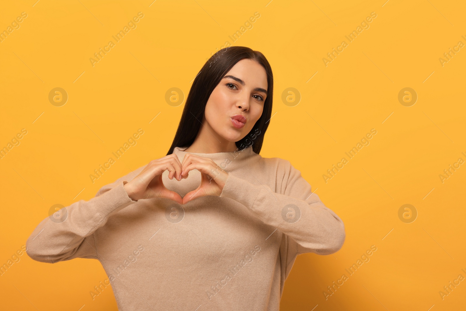 Beautiful young woman blowing kiss and making heart with hands on orange background Photo of Beautiful young woman blowing kiss and making heart with hands on orange background