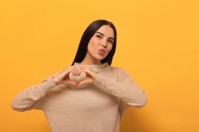 Photo of Beautiful young woman blowing kiss and making heart with hands on orange background