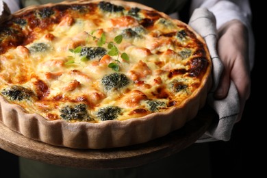 Photo of Woman holding wooden board with delicious homemade salmon quiche on black background, closeup