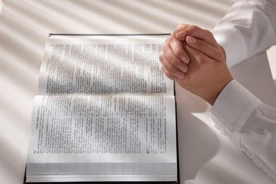 Photo of Woman holding hands clasped while praying over Bible at white table, closeup