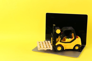 Photo of Laptop and toy forklift with wooden pallet on yellow background, space for text