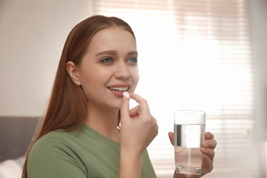 Photo of Young woman with glass of water taking vitamin pill at home