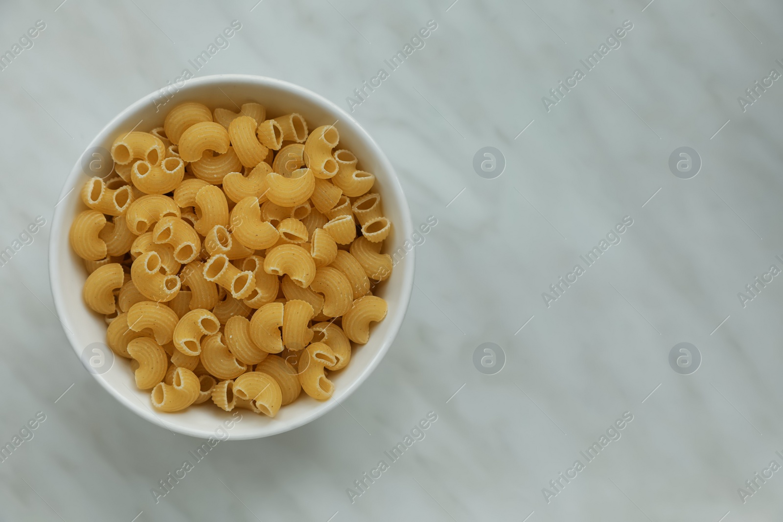 Photo of Bowl with uncooked horns pasta on white marble table, top view. Space for text