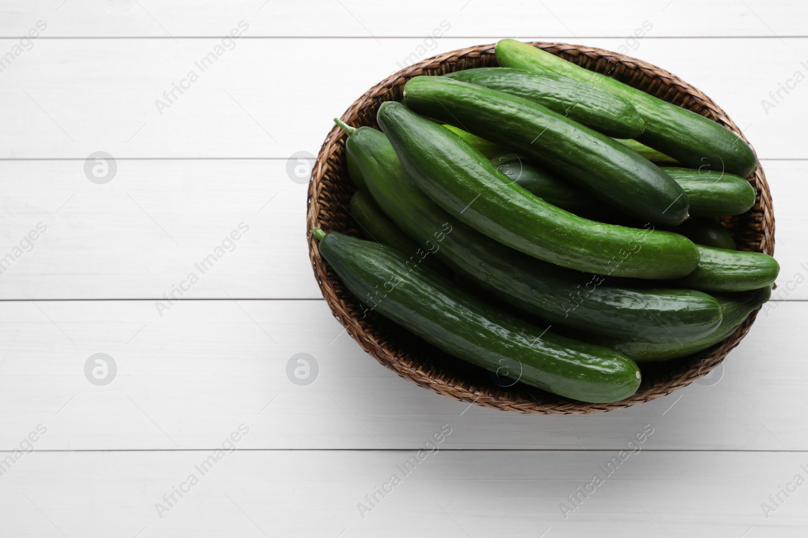 Photo of Fresh ripe cucumbers in wicker bowl on white wooden table, top view. Space for text