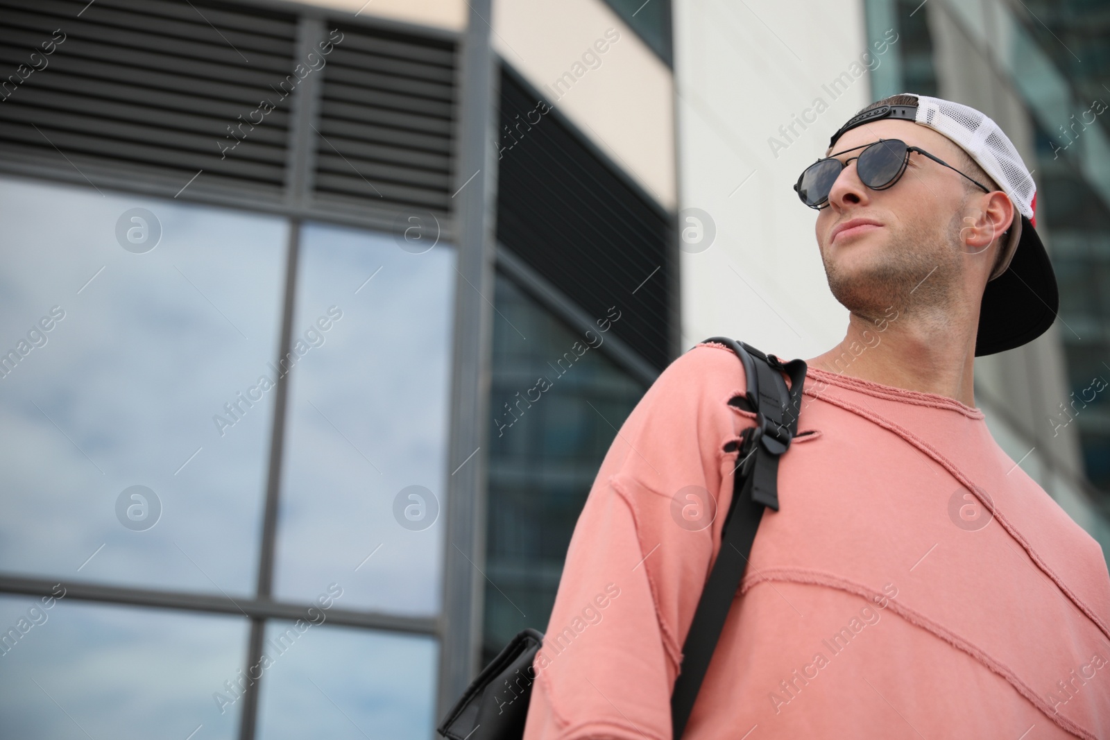 Photo of Handsome young man with stylish sunglasses and backpack near building outdoors, low angle view. Space for text