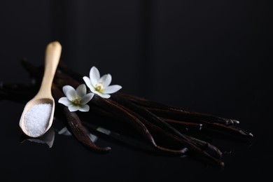 Photo of Vanilla pods, flowers and spoon with sugar on dark mirror table, closeup