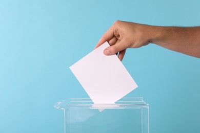 Man putting his vote into ballot box on light blue background, closeup