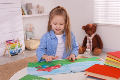 Photo of Cute little girl reading book at table in room