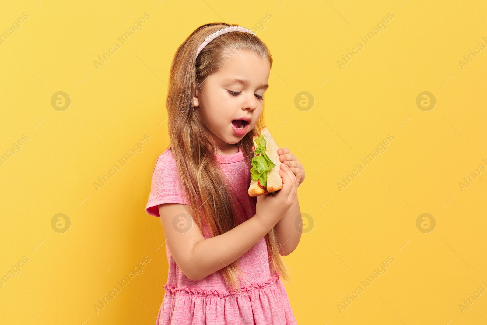 Photo of Cute little girl eating tasty sandwich on yellow background