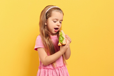 Cute little girl eating tasty sandwich on yellow background