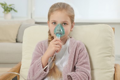 Photo of Little girl using nebulizer for inhalation in armchair at home