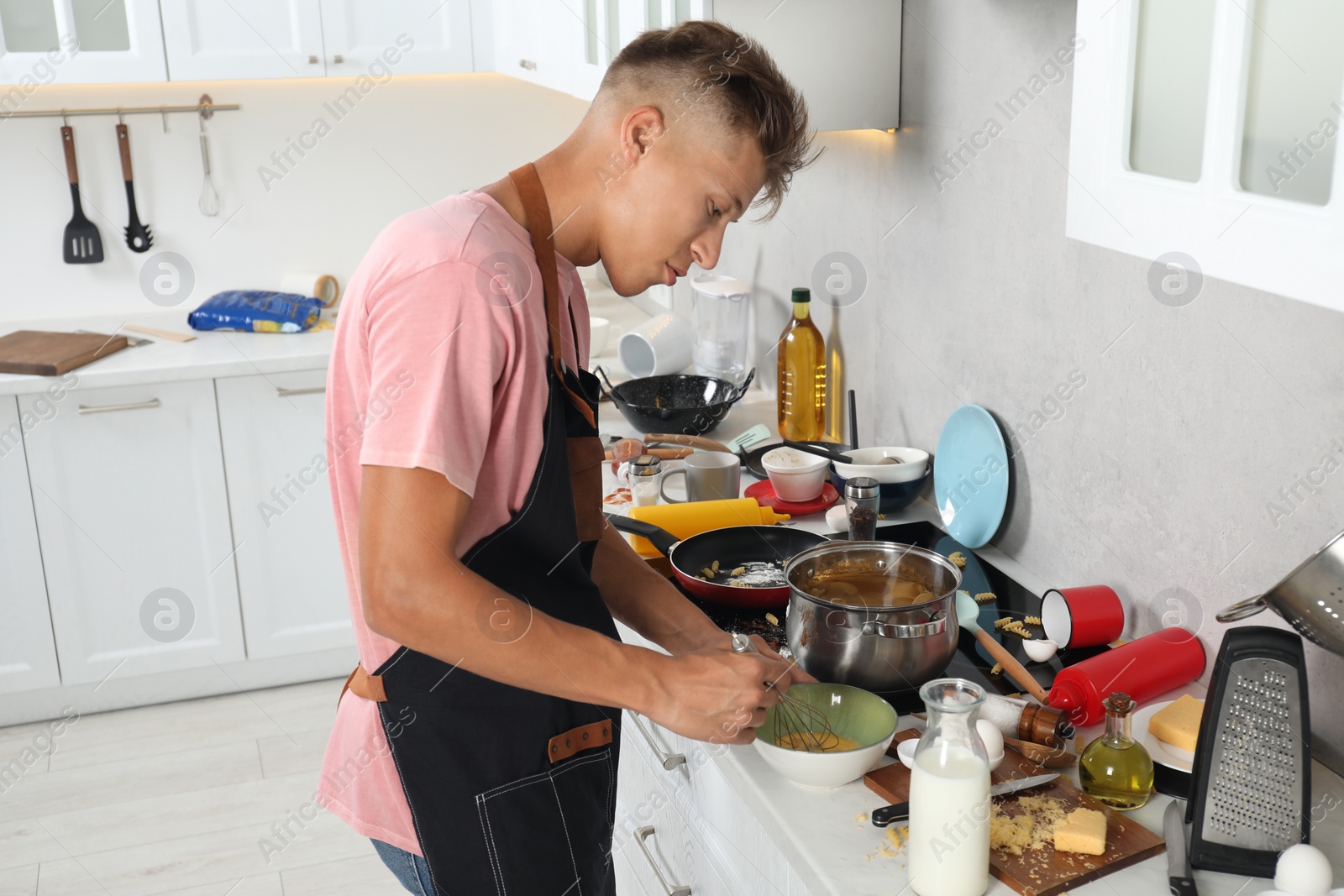 Photo of Man whisking eggs in bowl in messy kitchen. Many dirty dishware and utensils on stove and countertop