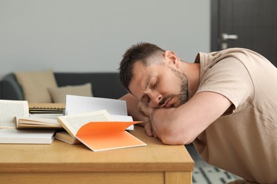 Photo of Tired man sleeping among books at wooden table indoors
