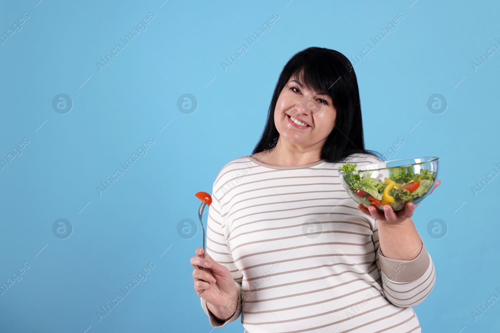 Photo of Beautiful overweight woman eating salad on light blue background, space for text. Healthy diet