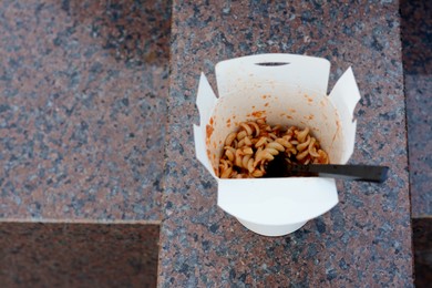 Photo of Paper box of takeaway noodles with fork on stone surface, above view and space for text. Street food