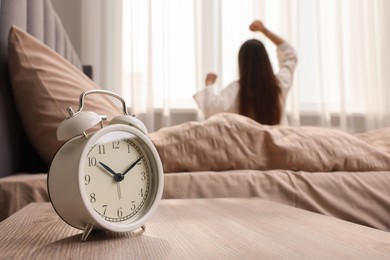 Photo of It's lazy morning o'clock. Alarm clock on bedside table and woman stretching in room, selective focus
