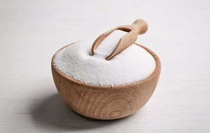 Wooden bowl with natural salt and scoop on white table, closeup