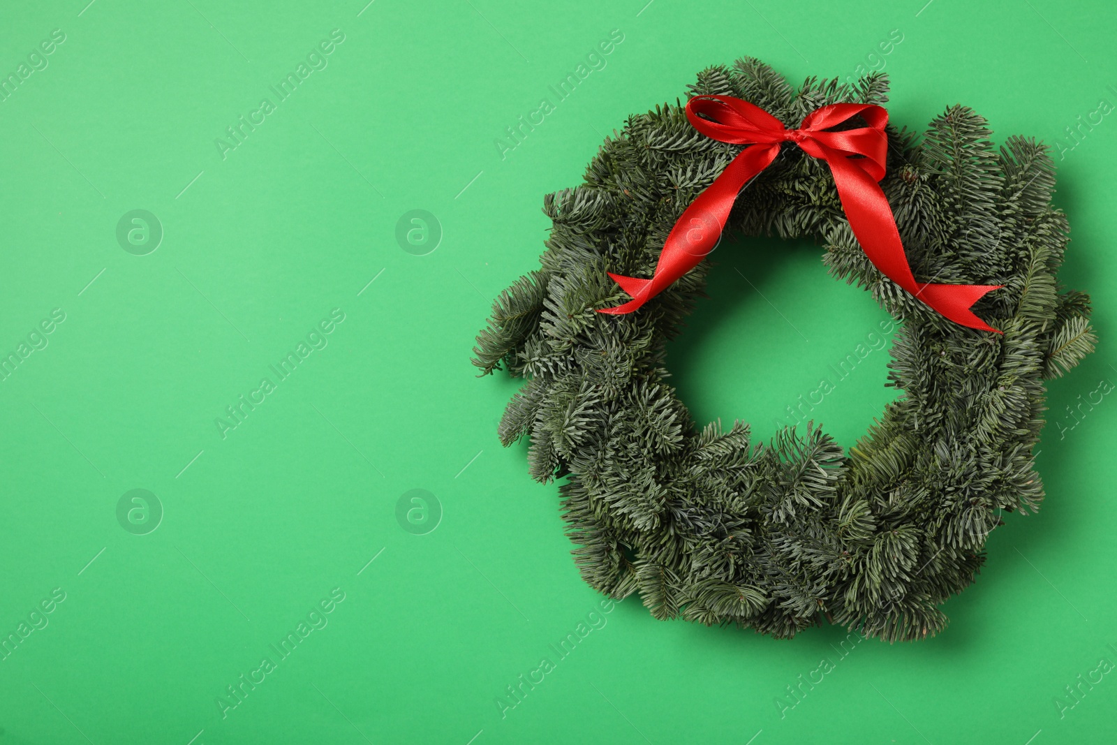 Photo of Christmas wreath made of fir tree branches with red ribbon on green background, space for text