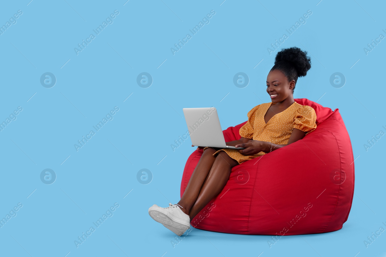 Photo of Beautiful young woman with laptop sitting on beanbag chair against light blue background. Space for text