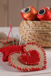 Photo of Heart of burlap fabric with red stitches, pincushion and needles on white wooden table, closeup. Space for text