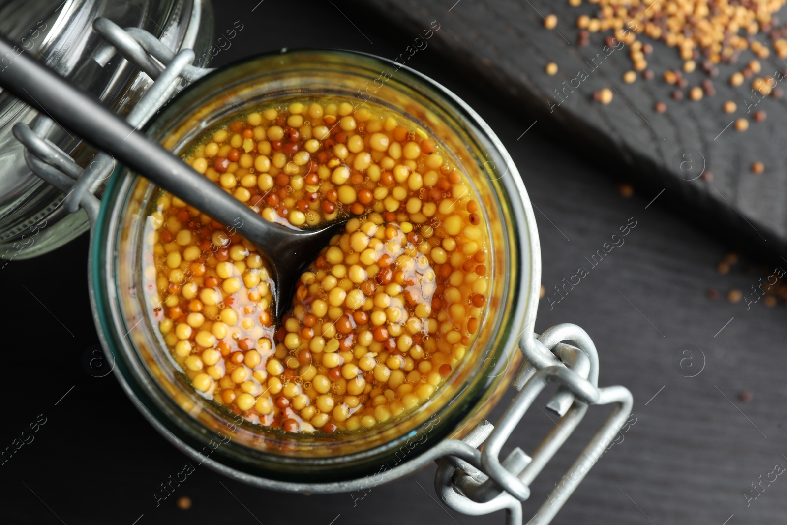 Photo of Whole grain mustard and spoon in jar on black wooden table, top view