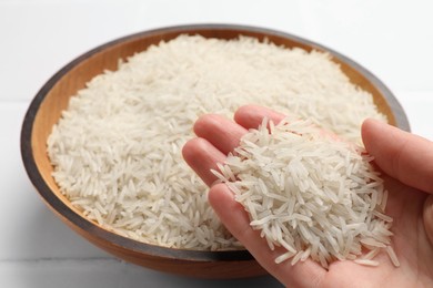 Photo of Woman holding raw basmati rice near bowl at white table, closeup