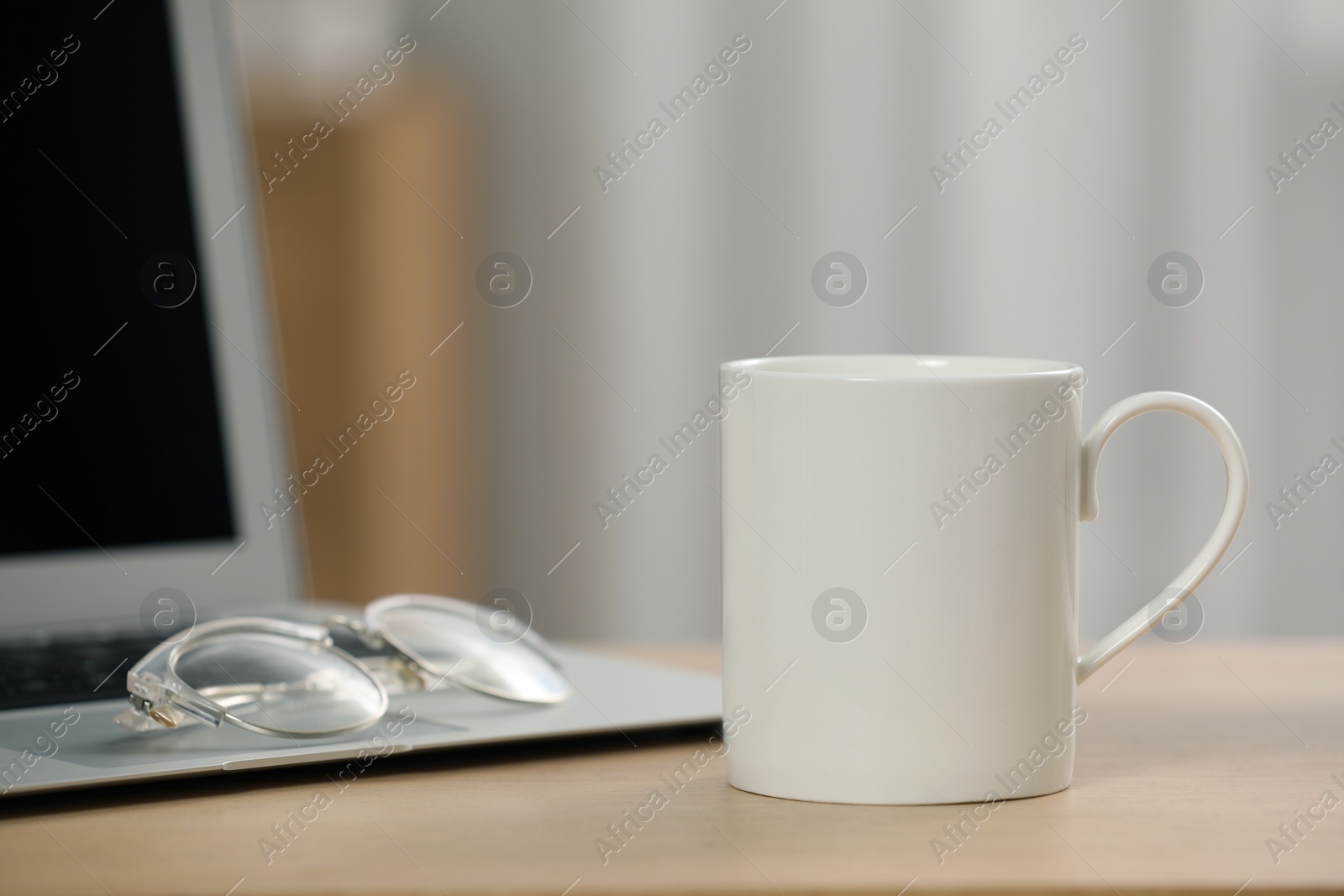 Photo of White ceramic mug, glasses and laptop on wooden table at workplace. Space for text