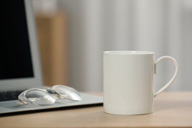 Photo of White ceramic mug, glasses and laptop on wooden table at workplace. Space for text